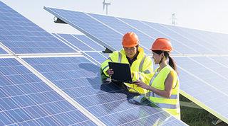 Two people in yellow hi-vis vests and orange safety helmets leaning on a solar panel in a field, looking at an iPad